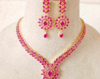 May include: A gold-toned jewelry set featuring a necklace and matching earrings. The necklace and earrings are adorned with vibrant pink gemstones arranged in a floral pattern. The set includes a pendant and dangling earrings.