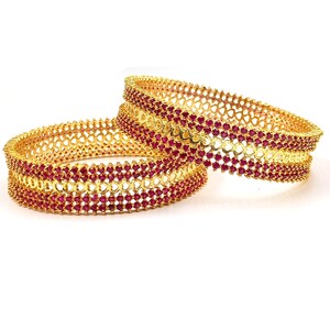 Peut inclure: Deux bracelets joncs dorés dotés d'une bande centrale de maillons métalliques ajourés en forme de cœur, bordée de deux rangées de petites pierres rondes rouge vif de chaque côté. Les bracelets circulaires ont une finition métallique polie.