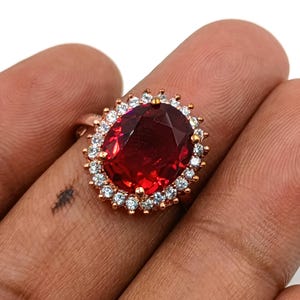 Pode incluir: Um anel com uma grande pedra preciosa vermelha escura de corte oval, montada numa banda de metal em tom de ouro rosa. A pedra central vermelha é rodeada por um halo de pequenas pedras brancas claras e cintilantes em engaste de garras.