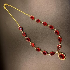May include: A gold-tone necklace featuring a series of teardrop-shaped red stones linked together. Each red stone is separated by a small clear accent stone. The necklace terminates in a larger teardrop-shaped pendant with a clear stone border.