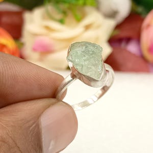 May include: A silver ring featuring a light green, raw-cut gemstone. The gemstone is irregularly shaped and set in a polished silver band. The ring is held by a hand, with a blurred floral background.