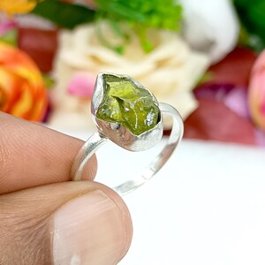 May include: A silver ring featuring a raw, olive-green peridot gemstone. The peridot is irregularly shaped and set in a silver bezel. The ring band is a simple, polished silver. The background is blurred with colorful flowers.