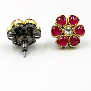 May include: A pair of flower-shaped stud earrings featuring six teardrop-shaped magenta stones surrounding a single clear center stone. The settings are gold-toned with a dark, oxidized metal backing and a standard post-style earring back.