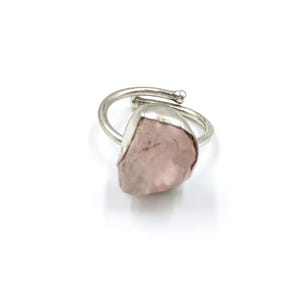 Peut inclure: Une bague argentée ornée d'une grosse pierre de quartz rose de forme irrégulière et de couleur rose pâle. L'anneau de la bague épouse le doigt, avec un petit détail décoratif en haut. La pierre est sertie dans une lunette argentée.