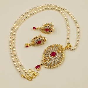May include: A jewellery set consisting of a single strand of white pearls and a gold-toned pendant featuring a central oval red stone surrounded by clear crystals. The set includes matching drop earrings with similar red stones and clear crystal accents.