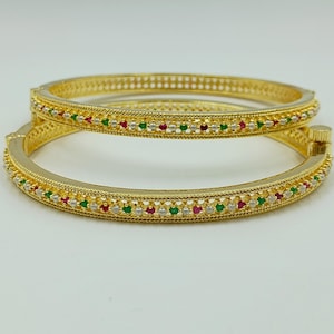 Könnte beinhalten: Ein Paar goldfarbene Klapparmreifen mit texturiertem Rand und einer zentralen Reihe aus abwechselnd kleinen, runden roten, grünen und weißen Steinen. Die Armreifen verfügen über einen sicheren Verschlussmechanismus und eine polierte Oberfläche.