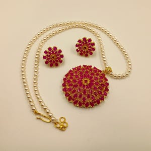 May include: A jewelry set consisting of a necklace and two stud earrings. The necklace features a strand of white pearls with a large, circular gold-tone pendant adorned with vibrant pink, marquise-cut stones arranged in a floral pattern.