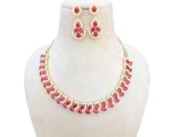 May include: A gold-toned necklace and earring set. The necklace and earrings feature ruby-red teardrop-shaped stones and small white stones. The jewelry is displayed on a mannequin head against a white background.