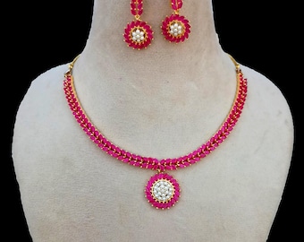 May include: A gold-toned necklace and earring set. The necklace and earrings feature a vibrant pink stone design with a central cluster of white stones. The earrings are a drop style, and the necklace has a matching pendant.