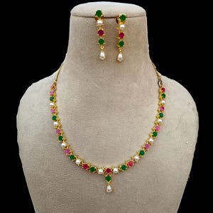 Puede incluir: Un conjunto de collar y pendientes dorados. El collar y los pendientes presentan un patrón repetido de piedras preciosas verdes, rosas y blancas. Los pendientes tienen una perla colgante. El collar tiene un solo colgante de perla.