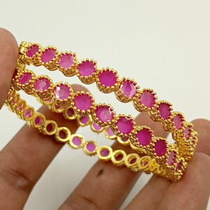 May include: A pair of gold-toned bangles featuring a continuous row of round, vibrant pink stones set in scalloped metal settings. The circular bracelets have a polished metallic finish and a uniform design, showcasing a bright, saturated color palette.