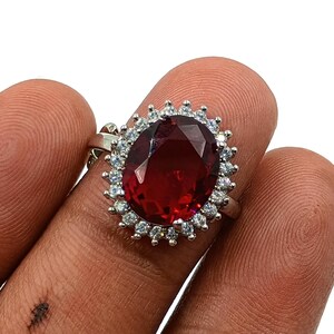 Puede incluir: Un anillo de tono plateado con una gran piedra preciosa de color rojo intenso y corte ovalado en el centro. La piedra central está rodeada por un halo de pequeñas piedras blancas transparentes de corte redondo en un marco de metal festoneado.