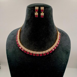 May include: A gold-toned necklace and matching drop earring set featuring oval-shaped deep pink stones bordered by small white beads. The necklace has a multi-layered design with a row of pink stones at the base and white beads above.