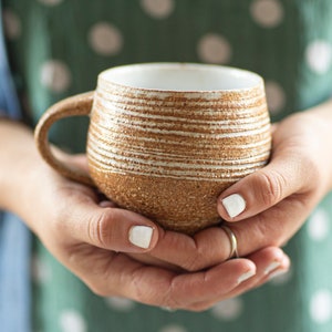 May include: A brown and white ceramic mug with a textured, striped design. The mug is being held in two hands.
