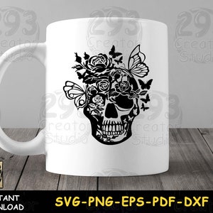 May include: White ceramic mug with a black silhouette of a skull with flowers and butterflies on the front. The mug is on a wooden surface.