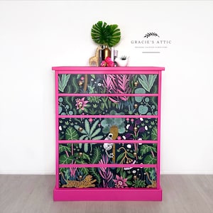 May include: A pink chest of drawers with five drawers. Each drawer is covered with a different patterned paper featuring tropical plants, animals, and flowers. The chest of drawers is labelled "Gracie's Attic Bespoke Painted Furniture".