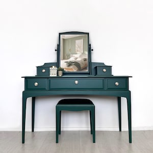 May include: A dark green vanity table with a matching stool, featuring a rectangular swivel mirror, three drawers with round silver knobs, and two small top storage compartments. The vanity has slender legs and a smooth, matte finish.