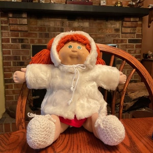 May include: A Cabbage Patch Kid doll wearing a white faux fur coat with a hood, red knit pants, and white crocheted booties. The doll has red yarn hair and green eyes.