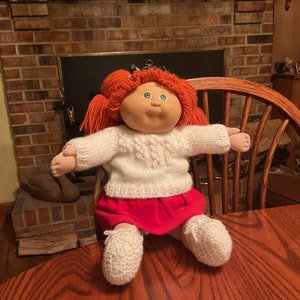 May include: A red-haired Cabbage Patch Kid doll wearing a white knitted sweater, a red skirt, and white knitted booties. The doll is sitting on a wooden chair in front of a brick fireplace.