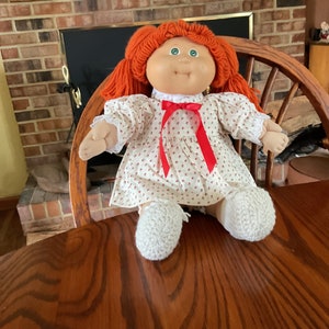 May include: A Cabbage Patch Kid doll with red yarn hair, green eyes, and a white dress with red polka dots. The doll is wearing white crocheted booties and is sitting on a wooden chair.