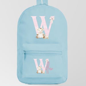 Può includere: Uno zaino azzurro con tasca frontale e maniglia superiore. Presenta una grande lettera rosa "W" con un'illustrazione di mucca e orsacchiotto. La parola "Willow" è scritta sotto.