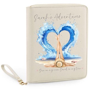 May include: Cream-colored travel wallet with a wrist strap. The front features a watercolor illustration of a person relaxing on a beach, with legs raised, framed by a heart-shaped wave. Text reads "Sarah's Adventures" and "Sun on my nose. Sand in my toes."