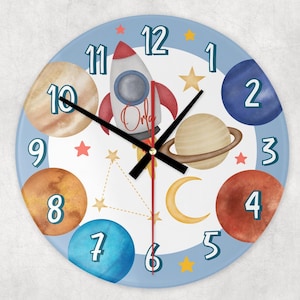 May include: A round wall clock with a light blue border and a white center featuring a rocket ship, planets, stars, and the name "Orla". The clock hands are black, and the numbers are in a playful font.