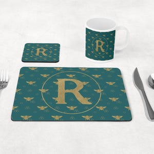 Könnte beinhalten: Ein tealfarbenes Platzset, ein Untersetzer und ein Tassenset mit einem goldenen Buchstaben "R"-Design. Das Platzset und die Tasse weisen ein sich wiederholendes Muster aus goldenen Bienen auf. Eine Gabel, ein Messer und ein Löffel befinden sich auf der rechten Seite des Bildes.