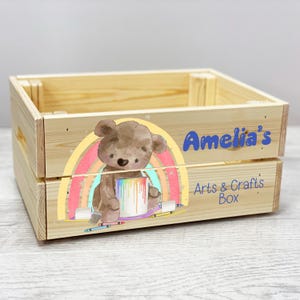 May include: A wooden crate with a rainbow and a teddy bear holding a paint can. The crate is labeled "Amelia's Arts & Crafts Box".