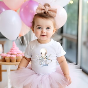 Puede incluir: Un bebé con una camiseta blanca con un pulpo y el nombre "Louise" en escritura azul. El bebé también lleva un tutú rosa y un gran lazo brillante. Globos y cupcakes se ven en el fondo.