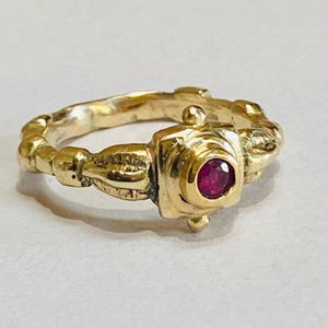May include: A gold ring featuring a red gemstone set in a square bezel. The band is textured and has a decorative design.