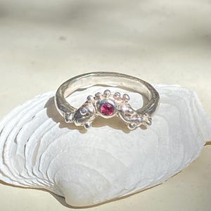 May include: A silver ring with a distinctive design, featuring a central pink gemstone and decorative side elements. The ring is presented on a white seashell, highlighting its intricate craftsmanship and elegant style. The ring's design is reminiscent of a crown.