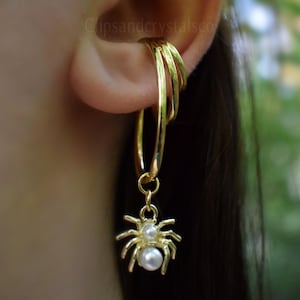 May include: Gold-toned ear cuff with three stacked hoops and a dangling spider charm. The spider has a detailed body and two faux pearl accents. The jewelry is worn on the ear, with a blurred green background.