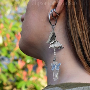 May include: Silver butterfly earrings with a clear crystal and chain detail. The earrings are dangling and have a layered design. The ear cuff is silver and wraps around the ear. The text "Clipsandcrystalsco" is visible.