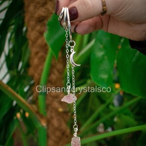 Könnte beinhalten: Ein silberfarbenes Pendel mit einem Mondsichel-Anhänger, zwei rosa Kristallen und einer Kette. Das Pendel hängt an einem silbernen Ring. Der Hintergrund ist ein sattes Grün mit dem Text "Clipsandcrystalsco".