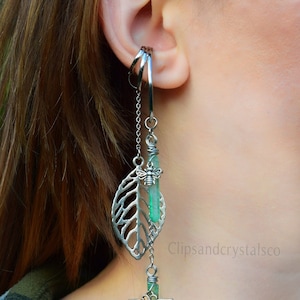 May include: Silver-toned ear cuff with a dangling earring featuring a leaf, bee, and dragonfly design. The earring includes a green crystal and chain accents. The brand name "Clipsandcrystalsco" is visible.