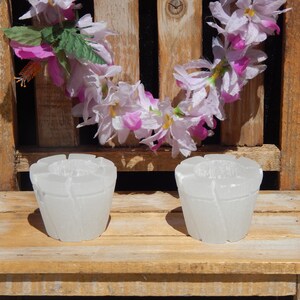 May include: Two white, textured votive candle holders with a floral design sit on a wooden surface. A garland of pink and white flowers hangs in the background, creating a decorative display.