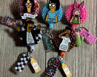 May include: Five badge reels featuring characters from Bob's Burgers. Each reel has a circular base covered in colorful rhinestones, a character charm with text, and a small dangling charm like a beer bottle or cactus. The characters include Linda, Tina, Louise, Bob, and Jimmy Pesto Jr.