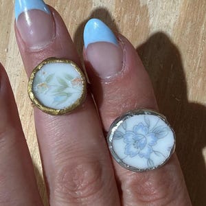May include: Two rings with floral designs. One ring has a gold-colored border, and the other has a silver-colored border. The floral designs feature blue, orange, and green accents on a white background. The rings are round.
