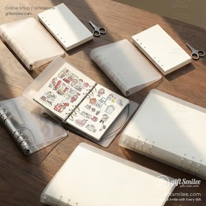 以下が含まれることがあります： 木製の表面に、透明なプラスチックカバーが付いた白いノートとバインダーの詰め合わせが配置されています。1冊のノートが開いており、カラフルなイラストが表示されています。小さな銀色のハサミも存在します。