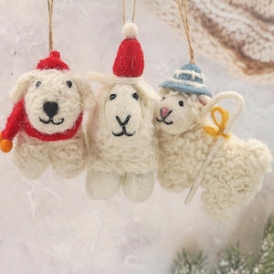 May include: Three woolly ornaments in the shape of sheep and a dog, each with a hat or scarf. The dog has a red scarf and hat. One sheep wears a red hat, and the other a blue hat, holding a yellow shepherd's crook. They are suspended by twine.