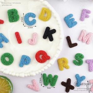 Może przedstawiać: Różne kolorowe litery z tkaniny, w tym niebieskie, zielone, żółte i różowe, ułożone na białym talerzu. Litery różnią się rozmiarem, niektóre mierzą 2,5 cm wysokości i 2,3 cm szerokości. Litery wykonane są z teksturowanego materiału.