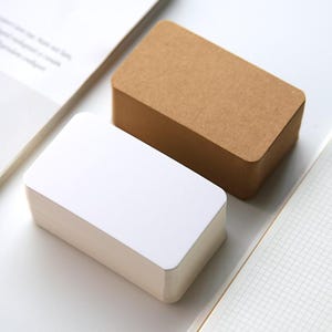 Peut inclure: Deux piles de cartes rectangulaires aux coins arrondis. Une pile est blanche, l'autre est marron. Les cartes sont sur une surface blanche, avec un carnet et des pages en arrière-plan.
