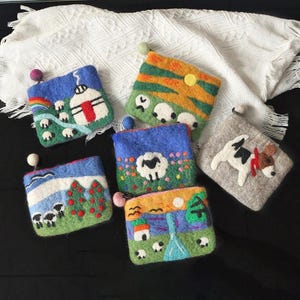 Puede incluir: Una colección de coloridas bolsas de lana afieltrada, cada una con una escena de paisaje única. Los diseños incluyen ovejas, una yurta, un arcoíris y una vaca. Las bolsas tienen un pequeño lazo y una bola decorativa en la parte superior.