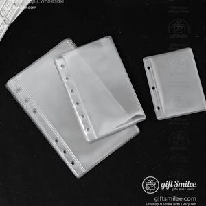 May include: Clear plastic binder sleeves in various sizes. The sleeves have multiple holes for insertion into a binder. The image shows three sleeves, two larger and one smaller, arranged on a dark surface. The sleeves are transparent.