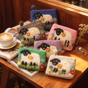 May include: A collection of colorful, handcrafted wool coin purses. Each purse features a white sheep with a black face and legs, set against a background of blue, brown, pink, purple, green, and cream. Floral and grass details surround the sheep.