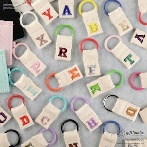 Puede incluir: Una colección de llaveros de lona con letras bordadas en varios colores. Cada llavero tiene un anillo colorido y una etiqueta rectangular de lona con una sola letra. Las letras son de diferentes colores y los anillos son de varios colores.