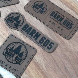 May include: Four brown leather patches with black laser-engraved text and images. The patches feature the text "BARN 605" and "BLACK HILLS, SOUTH DAKOTA". The patches are available in two sizes: 4.25 inches by 2 inches and 2.25 inches by 2.25 inches.