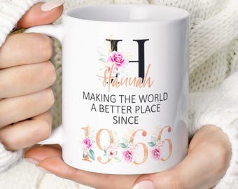 Może przedstawiać: Biały ceramiczny kubek z dużą czarną literą H, imieniem Hannah zapisanym różowozłotą kursywą oraz czarnym napisem MAKING THE WORLD A BETTER PLACE SINCE 1966. Wzór zawiera różowe kwiatowe akcenty wokół litery H oraz daty 1966.