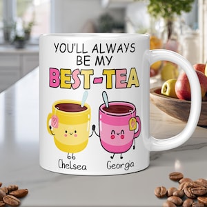 White personalised mug with the text "You'll always be my best-tea" and two cartoon teacups holding hands in a kitchen worktop with a kitchen on the background.